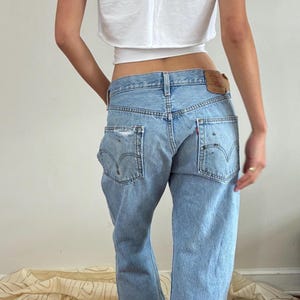 May include: A white short-sleeved cropped t-shirt paired with light-wash blue denim jeans. The jeans feature a classic five-pocket design, a brown leather patch on the back waistband, visible stitching on the back pockets, and subtle distressing.