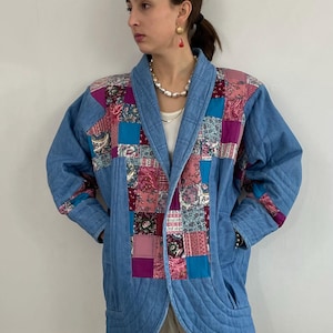 May include: A quilted denim jacket with a patchwork design. The jacket features a light blue denim base with a colorful patchwork of pink, purple, and teal fabrics. The jacket has a shawl collar and two front pockets.