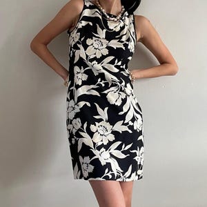 May include: A sleeveless knee-length dress featuring a bold black background with a large-scale white floral pattern. The garment has a high, rounded neckline and a straight, shift-style silhouette that falls to just above the knees.