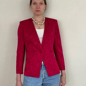 May include: A magenta blazer with a subtle floral pattern. The blazer has a single-button closure and three-quarter sleeves. The model is wearing a white top, blue jeans, and a pearl necklace. A gold bracelet is visible on the wrist.