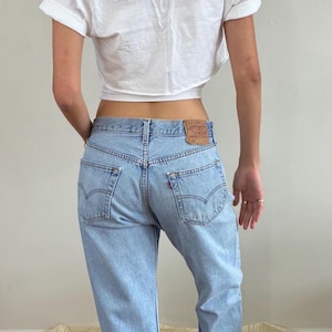 May include: A white short-sleeved t-shirt with a crew neckline and rolled sleeves paired with light-wash blue denim jeans. The jeans feature a tan leather patch on the back waistband, two back pockets with stitched detailing, and a red tab.