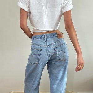 May include: A white short-sleeved t-shirt with rolled sleeves and a cropped hem paired with light blue denim jeans. The jeans feature a tan leather patch on the back waistband, two back pockets with arcuate stitching, and a small red brand tab.
