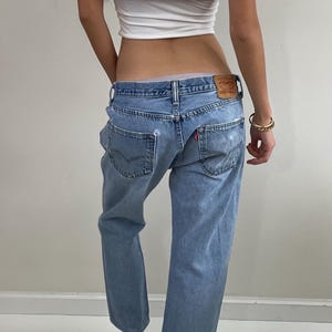 May include: Light blue denim jeans with a distressed finish, worn with a white crop top and burgundy mules. The jeans feature a classic five-pocket design and a leather patch. A gold bracelet is visible.