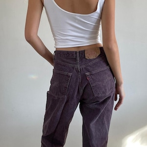 Könnte beinhalten: Ein weißes Tanktop kombiniert mit lila Jeans. Die Jeans haben ein klassisches Fünf-Taschen-Design und ein verwaschenes, strukturiertes Aussehen. Ein kleiner Lederaufnäher ist auf der Rückseite der Jeans zu sehen.