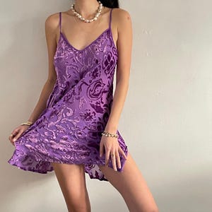 May include: A purple slip dress with a floral pattern. The dress has thin straps and a V-neckline. The fabric appears to be a soft, silky material. The dress is knee-length.