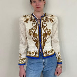 May include: Ivory jacket with ornate gold embroidery and blue trim. The jacket features a floral pattern and long sleeves. The jacket is worn with blue jeans. A pearl necklace and gold jewelry complete the look.