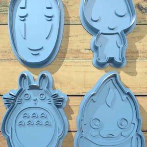 May include: Four light blue cookie cutters in various shapes. The shapes include a face with no mouth, a small figure with closed eyes, a round character with a belly, and a flame-shaped character. The cutters are made of plastic and have a silver metal outline.