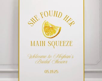May include: A white sign with a gold border and text. The text reads "SHE FOUND HER MAIN SQUEEZE" with a watercolor lemon slice illustration. Below, it says "Welcome to Meghan's Bridal Shower" and the date "05.19.25".