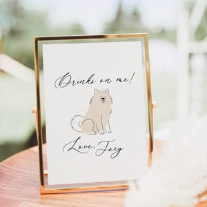 May include: A framed sign with a gold border and a white background. The sign features the text "Drinks on me!" above a cartoon illustration of a light brown dog. Below the dog is the text "Love, Joey."