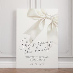 Può includere: Un cartello bianco con un grande ed elegante fiocco bianco nell'angolo in alto a destra. Il cartello recita "She's tying the knot!" e include il testo "Welcome to Meghan's Bridal Shower" e la data "08.18.2026".
