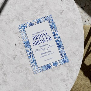 Flat lay of a blue and white bridal shower invitation with tile border on a marble tabletop, sunlight casting artistic shadows.