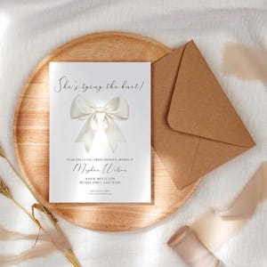 Può includere: Un invito per un addio al nubilato con il testo "She's tying the knot!" e una grande grafica a fiocco bianco. L'invito è su un vassoio di legno con una busta marrone. L'invito include il nome di Meghan Wilson e i dettagli dell'evento.