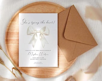Könnte beinhalten: Eine Brautparty-Einladung mit dem Text "She's tying the knot!" und einer großen weißen Schleifengrafik. Die Einladung liegt auf einem Holztablett mit einem braunen Umschlag. Die Einladung enthält den Namen Meghan Wilson und Veranstaltungsdetails.