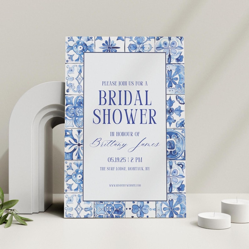 Blue and white tile-themed bridal shower invitation standing upright on a clean surface with modern white decor and tea light candles.