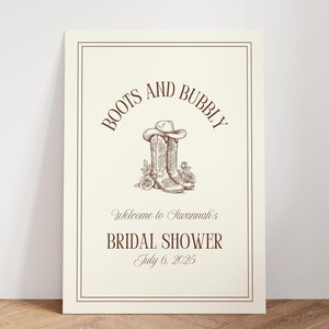 Puede incluir: Un letrero color crema con borde y texto marrón. Las palabras "Boots and Bubbly" forman un arco en la parte superior, sobre una ilustración de botas de vaquero y un sombrero. Debajo, se lee "Welcome to Savannah's Bridal Shower July 6, 2025."