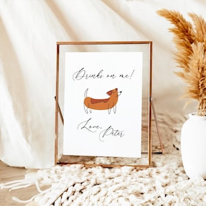 May include: A framed print with a brown dachshund illustration. The print has the text "Drinks on me!" and "Love, Peter" in elegant script. The frame is a light brown color and is displayed on a white surface.