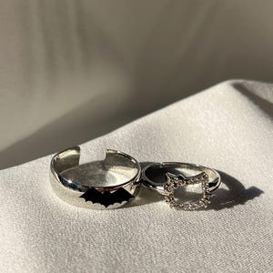 Puede incluir: Dos anillos de plata. Un anillo presenta una silueta de murciélago negro. El otro anillo es un diseño de Hello Kitty con diamantes de imitación transparentes.