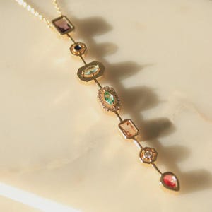 Puede incluir: Un collar de cadena de oro con una variedad de piedras preciosas de colores. Las piedras están engastadas en oro y están dispuestas en una línea vertical. El collar está sobre una superficie de mármol blanco.