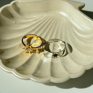 Può includere: Due anelli d'oro con un design a stella. Gli anelli sono su un piatto bianco a forma di conchiglia.