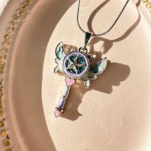 Może przedstawiać: Srebrny naszyjnik z zawieszką w kształcie różdżki z skrzydłami. Różdżka ma różowe serce, purpurowo-biały pasek i niebieską gwiazdę z turkusowym środkiem.