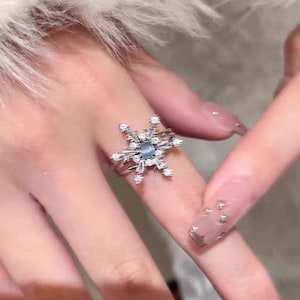 Puede incluir: Un anillo plateado con un diseño de copo de nieve. El anillo tiene una piedra central azul claro rodeada de piedras claras más pequeñas. El diseño del copo de nieve es detallado e intrincado, con múltiples puntas que se extienden hacia afuera. El anillo se lleva en un dedo.
