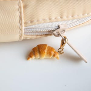 Könnte beinhalten: Eine beige Reißverschlusstasche mit einem kleinen Croissant-Anhänger am Reißverschluss.