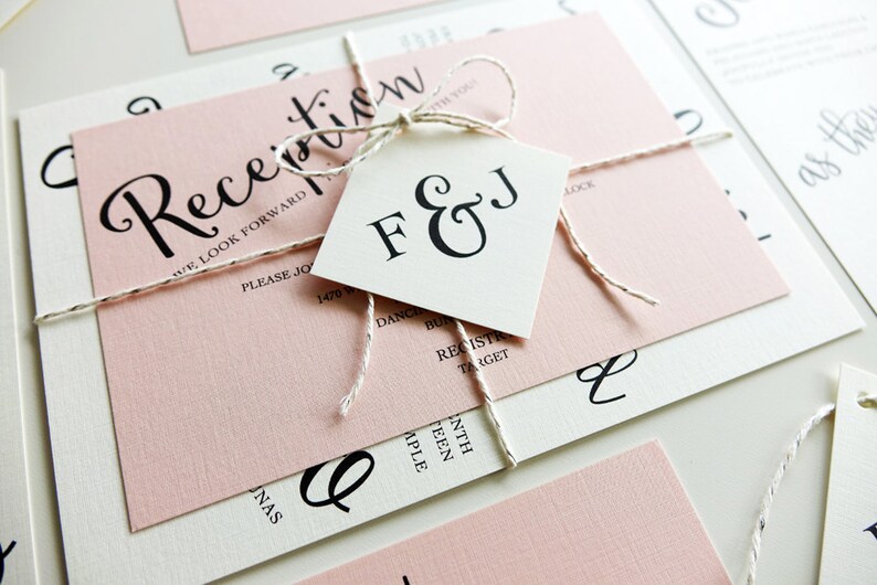 Peut inclure: Une invitation de mariage rose et blanche avec une &eacute;tiquette blanche attach&eacute;e avec de la ficelle. L'&eacute;tiquette porte les initiales "F & J" en &eacute;criture cursive noire. L'invitation dit "R&eacute;ception" en &eacute;criture cursive noire et "Nous avons h&acirc;te de c&eacute;l&eacute;brer avec vous".