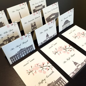 May include: Place cards with a white background and black and grey illustrations of famous buildings. The cards have pink cherry blossom accents and are printed with names in black cursive.