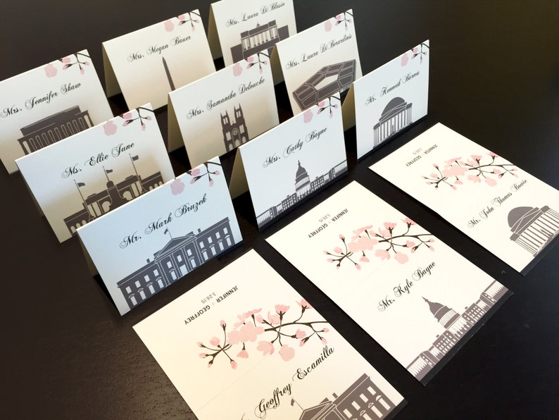 May include: Place cards with a white background and black and grey illustrations of famous buildings. The cards have pink cherry blossom accents and are printed with names in black cursive.