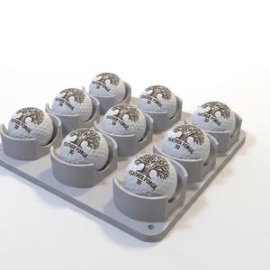 May include: A gray golf ball holder with nine golf balls. Each white golf ball has the text "FEATHER FORGE 3D" and a tree logo. The holder is gray and has individual slots for each ball.