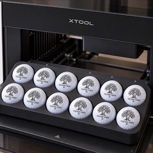 May include: A white and black XTool laser machine with an open front tray holding twelve white golf balls. Each golf ball features a black printed tree logo and the text "FEATHER FORGE 3D". A black box nearby reads "CUSTOM GOLF BALLS MADE EASY".