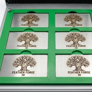 May include: A metal and green storage case with six rectangular compartments. Each compartment holds a silver plate with a detailed tree design and the text "FEATHER FORGE 3D".