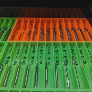 May include: A drawer organizer with green and orange compartments holding various drill bits. The bits are labeled with sizes and specifications, including "HSS" and "CB". The arrangement suggests organization for a workshop or tool storage.