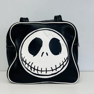 May include: Black handbag featuring a white Jack Skellington face design. The bag has a rounded shape with white trim and handles. The design is a graphic illustration of the character's face from the movie "The Nightmare Before Christmas".