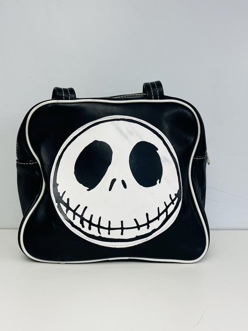 May include: Black handbag featuring a white Jack Skellington face design. The bag has a rounded shape with white trim and handles. The design is a graphic illustration of the character's face from the movie "The Nightmare Before Christmas".