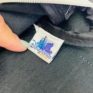May include: Close-up of a black bag with a white zipper and stitching. A tag with the Disneyland Resort logo is visible. The tag features a castle and bear graphic in blue and purple, with the words "Disneyland Resort" in orange.