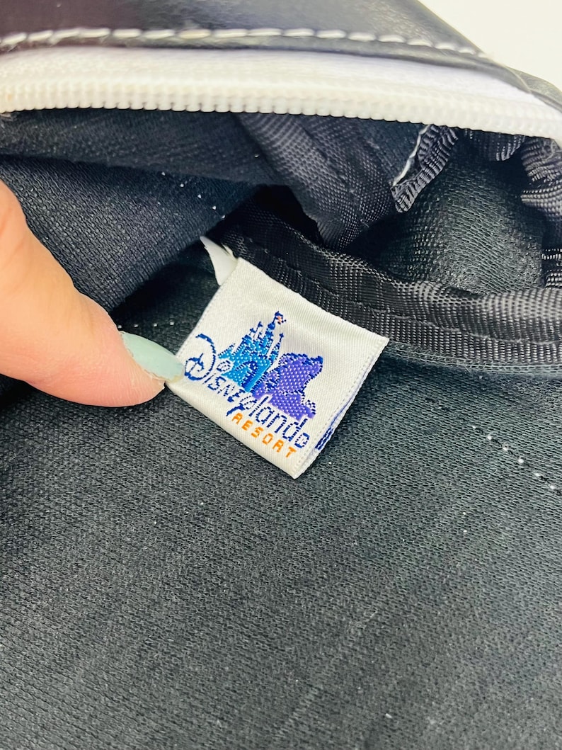 May include: Close-up of a black bag with a white zipper and stitching. A tag with the Disneyland Resort logo is visible. The tag features a castle and bear graphic in blue and purple, with the words "Disneyland Resort" in orange.