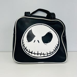 May include: Black square backpack featuring a white Jack Skellington face design. The bag has a top handle and white trim. The design is a graphic illustration of the character's face from the movie "The Nightmare Before Christmas".