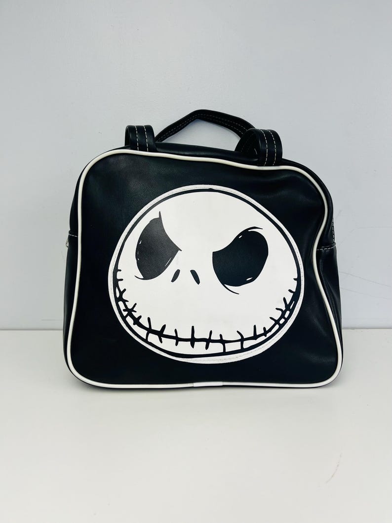 May include: Black square backpack featuring a white Jack Skellington face design. The bag has a top handle and white trim. The design is a graphic illustration of the character's face from the movie "The Nightmare Before Christmas".