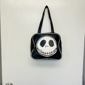 May include: Black tote bag featuring a white Jack Skellington face design. The bag has a square shape with a white trim and black handles. The bag is hanging on a white wall.