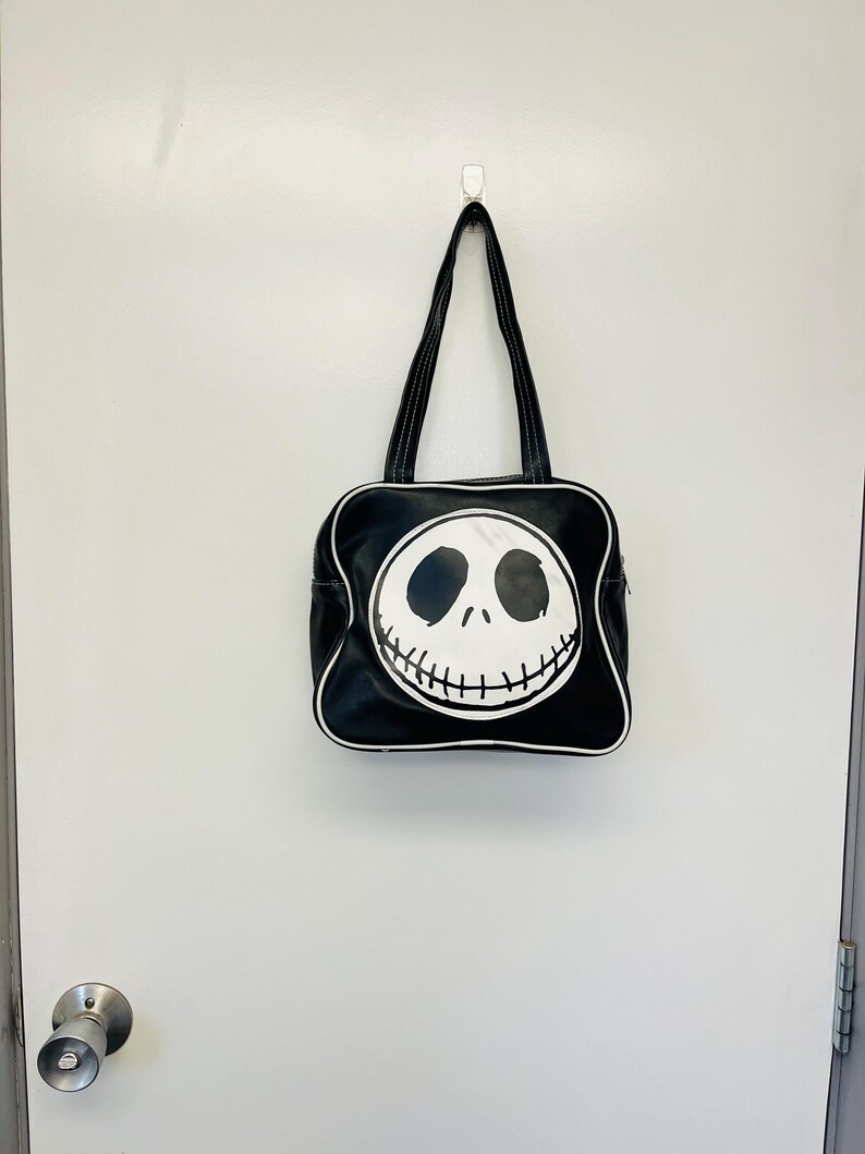 May include: Black tote bag featuring a white Jack Skellington face design. The bag has a square shape with a white trim and black handles. The bag is hanging on a white wall.