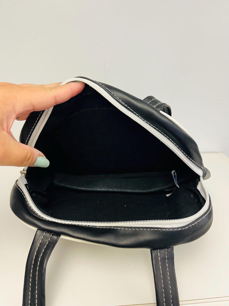 May include: Black and white handbag with a zippered top and two handles. The bag has white stitching and a white trim. The interior is black.