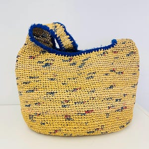 May include: A woven straw tote bag with a rounded shape. The bag is primarily yellow with small flecks of red, blue, and green. The top edge and handle are trimmed with a dark blue material. The bag is sitting on a white surface.