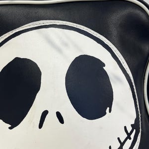 May include: Black and white satchel bag featuring a large graphic of Jack Skellington's face. The bag has a black exterior with a white outline and handle. The design includes large black eye sockets, a small nose, and a stitched grin.