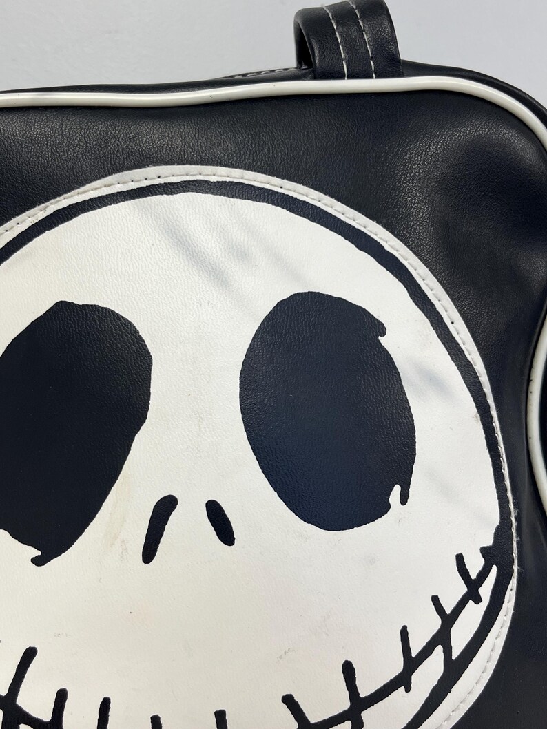 May include: Black and white satchel bag featuring a large graphic of Jack Skellington's face. The bag has a black exterior with a white outline and handle. The design includes large black eye sockets, a small nose, and a stitched grin.