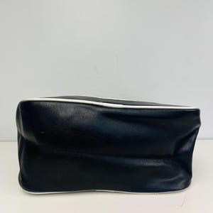 May include: A black handbag with a white trim. The bag is rectangular with a slightly rounded top and bottom. The bag is made of a smooth material and appears to be empty.