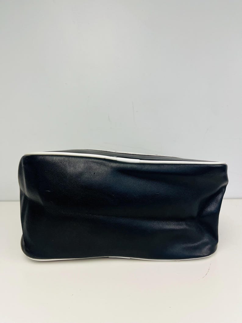 May include: A black handbag with a white trim. The bag is rectangular with a slightly rounded top and bottom. The bag is made of a smooth material and appears to be empty.