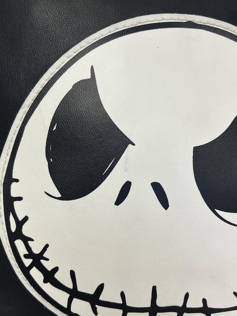 May include: Close-up of a black and white graphic design featuring a character's face. The face is white with black eyes and a stitched-like mouth. The background is black.