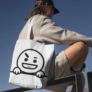 Puede incluir: Bolso tote blanco con una cara de dibujos animados en blanco y negro que expresa enfado. El bolso tiene un dise&ntilde;o sencillo con un contorno negro. Lo lleva una persona que viste una sudadera y unos pantalones cortos grises.