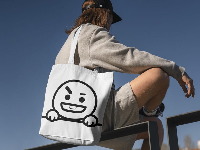 Puede incluir: Bolso tote blanco con una cara de dibujos animados en blanco y negro que expresa enfado. El bolso tiene un dise&ntilde;o sencillo con un contorno negro. Lo lleva una persona que viste una sudadera y unos pantalones cortos grises.